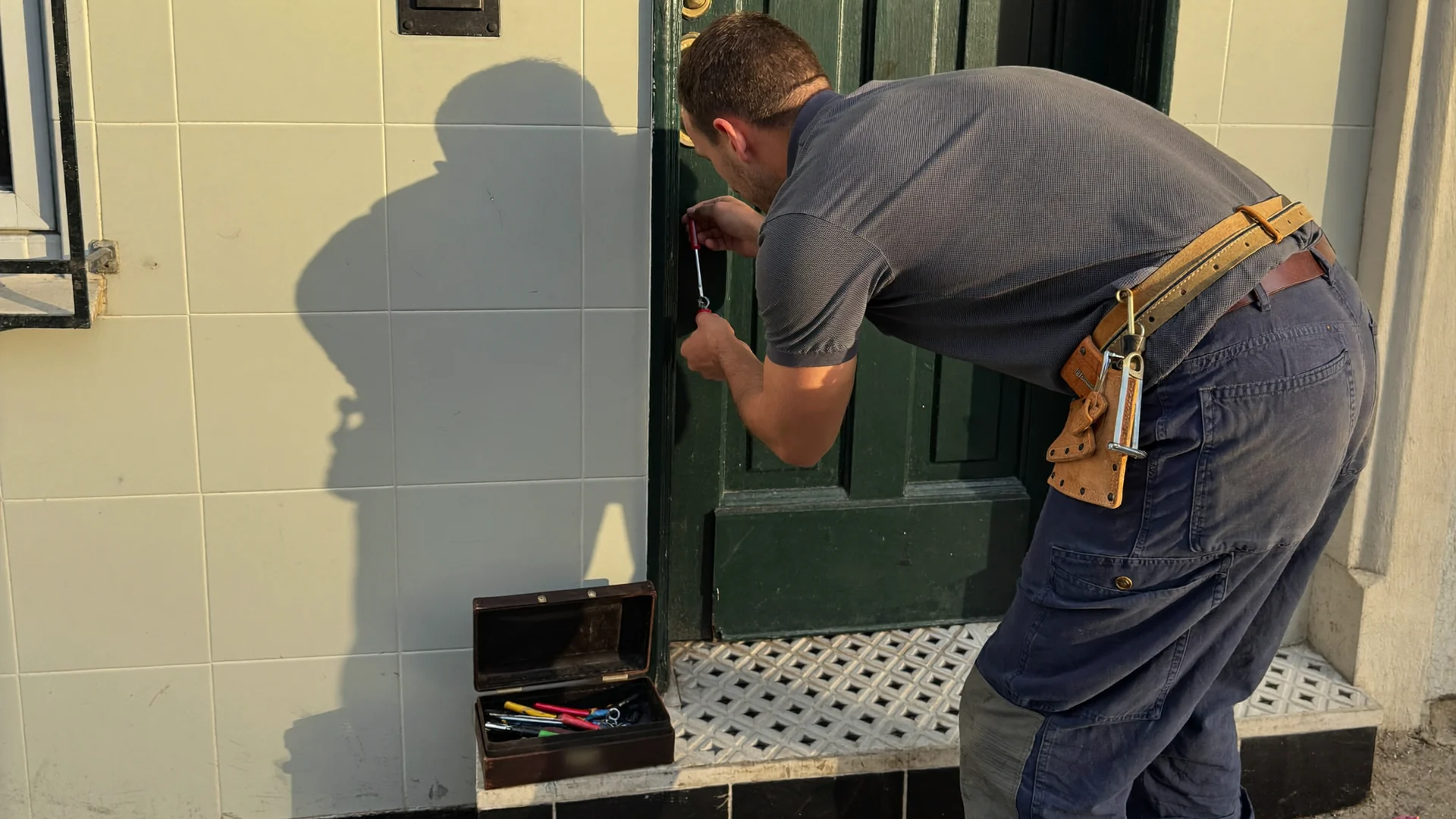 Locksmith in work clothing bent over the cylinder lock of a green front door in a sunlit tiled Lisbon doorway, tool case open on the step